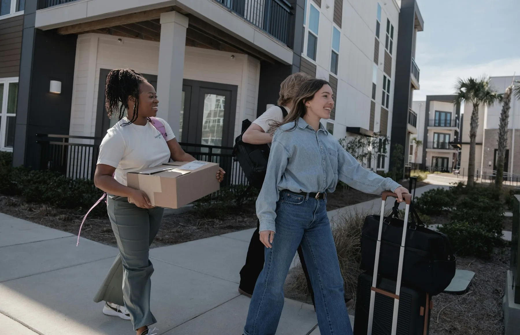 Young people carrying boxes and luggage toward a palm-lined apartment building